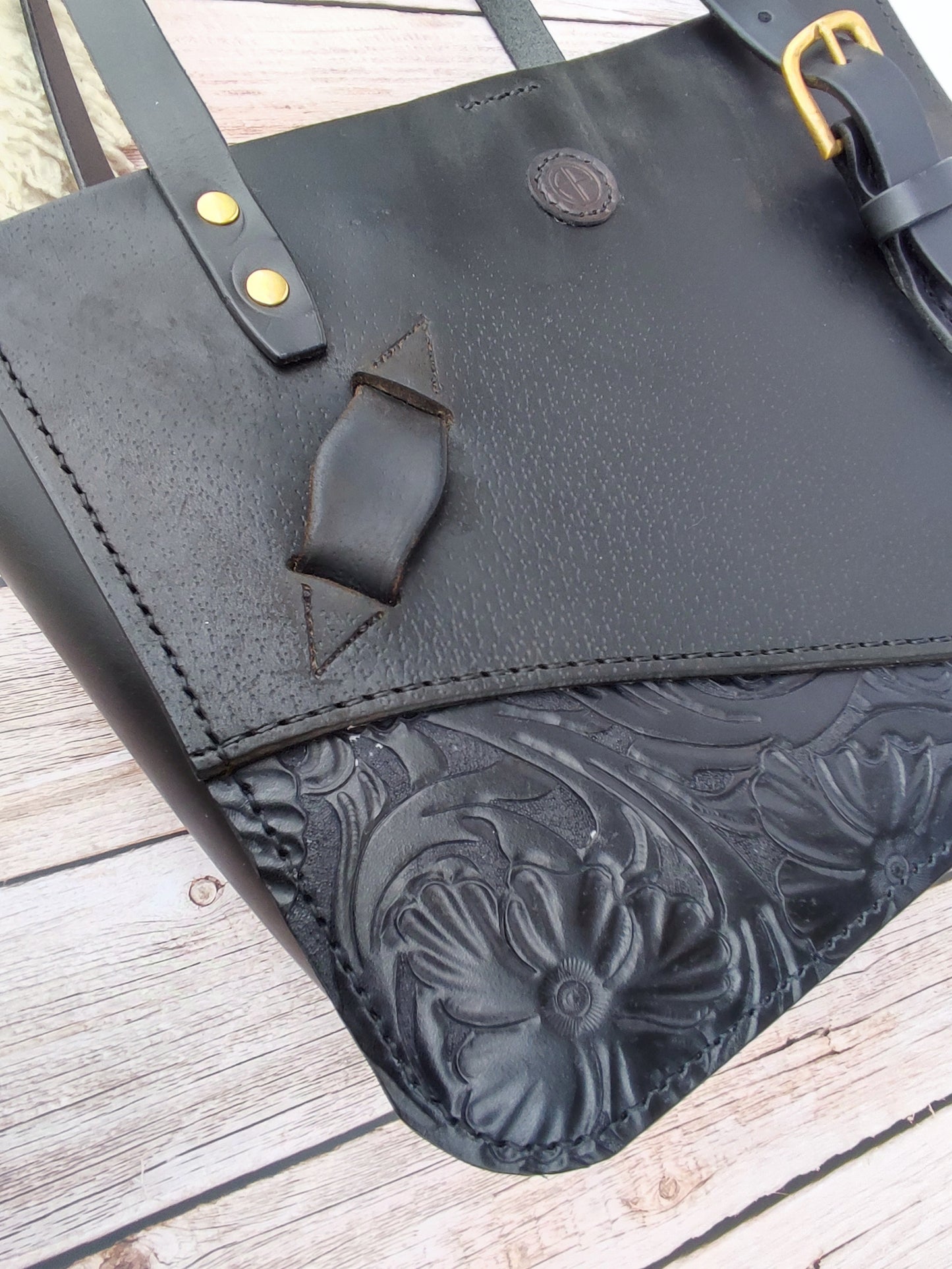 Détails du sac cabas noir qui le rend unique avec son passant d'étrivière et le morceau de cuir repoussé à la main au motif floral comme sur les selles westerns.